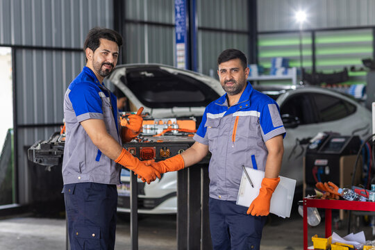 Two man automotive technicians shake hands beside electric vehicle battery during EV maintenance, teamwork, professional service, cooperation, and skilled automotive engineering.