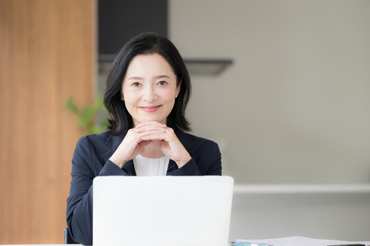 ノートパソコンで仕事をする女性会社員、オフィスで笑顔で働く顎に手を当てるビジネスウーマンのデスクワークカメラ目線