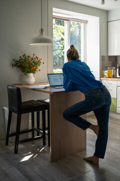 Woman works on laptop from home standing in unhealthy working position at high kitchen table, female freelancer leaning on desk in uncomfortable bad posture position causing harm to spine and back.