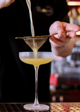 Pouring cocktail. Cold alcohol drink. Professional male barkeeper straining yellow aperitif using metal sieve preparing on counter.