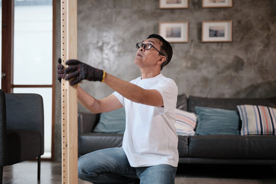 Asian mature man self-assembling flat-pack furniture with DIY tools, checking accuracy and strength of wooden parts, home improvement, craft installation, hobby workshop, handyman active lifestyle.
