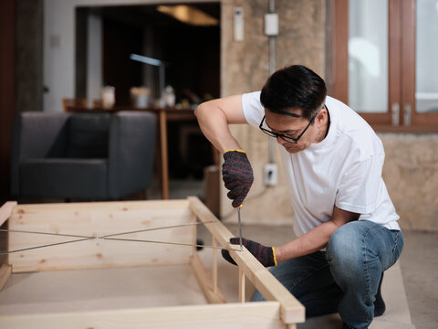 Asian mature man concentrates on self-assembling wooden furniture with DIY tools, tightening strength with a screwdriver, home improvement, hobby carpentry workshop, expert handyman, active lifestyle.