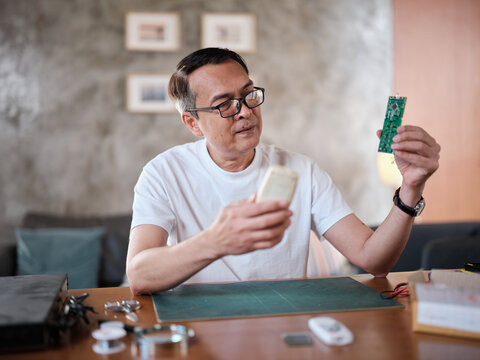 Mature Asian man checks and compares spare parts parcel, air conditioner remote control circuit board replacement, home electrical appliance fix, electronics component hobbyist, enthusiast workshop.