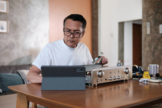 Mature Asian man disassembling audio equipment and self-learning, searching for manual guide to fix circuit board in tablet, home appliance DIY, electronics component hobby, handyman skill workshop.