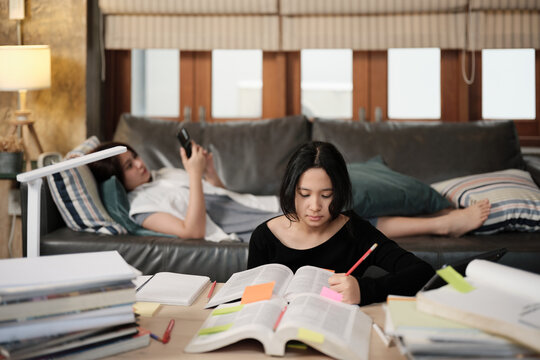 Two Asian teenage tutorials, girl concentrates on learning class at home with pile of homework, her friend uses mobile phone on sofa, self-study with textbooks, homeschooling together lifestyle.
