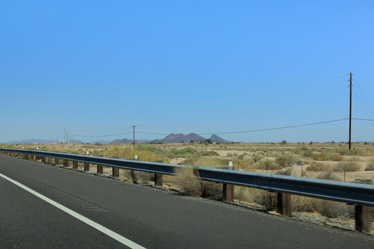Asphalt highway with a steel guardrail runs alongside a vast desert landscape featuring power lines and distant mountains under blue sky, Interstate-8 near Wellton, Arizona