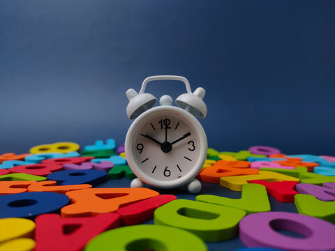 A small white twin-bell alarm clock stands centered amidst a scattered pile of colorful wooden letters on dark blue