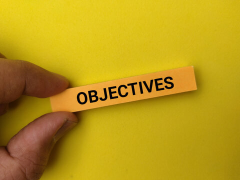 A hand holds a yellow paper strip printed with the word OBJECTIVES against a solid bright yellow background