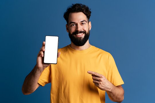 Cheerful bearded man in yellow tshirt pointing at a blank smartphone screen against a blue background