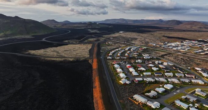 High oblique drone view over Grindavik, Iceland, showing a residential subdivision beside a long protective berm, black lava fields and barren volcanic terrain stretching toward distant hills.