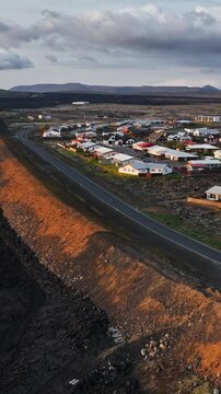 Vertical drone view of Grindavik, Iceland, showing a sunlit protective berm separating black lava terrain from a road and residential district beneath dramatic clouds.