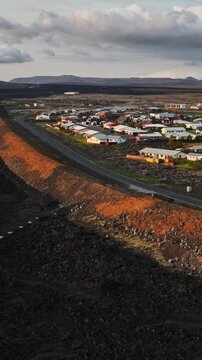 Vertical drone view of Grindavik, Iceland, showing a protective earth berm separating a vast black lava field from roads and homes on the volcanic town outskirts.