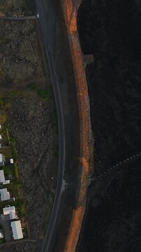 Vertical top-down drone view in Grindavik, Iceland, showing a curved road beside an orange protective berm separating nearby homes from a vast black lava field.