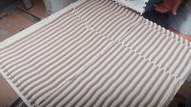 Close-up of a worker combing tile adhesive with a notched trowel, creating uniform ridges across a large ceramic slab during professional indoor renovation work.