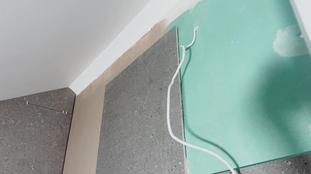 Close-up of a partially tiled interior wall with large gray ceramic panels, exposed electrical cable and green backing board during renovation and utility preparation.