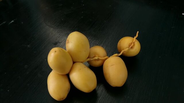 Close-up view of a bunch of fresh, young yellow dates (khalal stage) attached to a stem, resting on a textured dark black wooden table. Represents healthy raw food, Middle Eastern agricultural produce