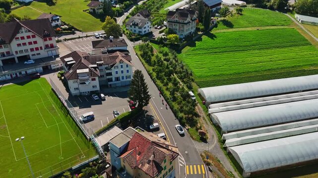Aerial view over Schwyz, Switzerland, showing greenhouse tunnels, farmland, homes, a football pitch and local roads across a green Alpine settlement on a sunny summer day.