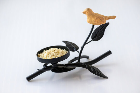 Decorative black metal branch stand with carved wooden bird and small dish filled with raw rice grains, isolated on white background.