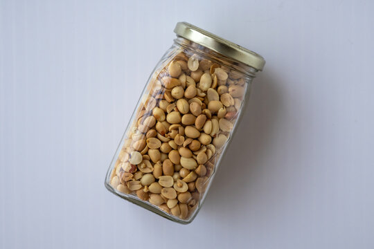 High angle view of a transparent glass jar filled with roasted peeled peanuts isolated on a plain light background.