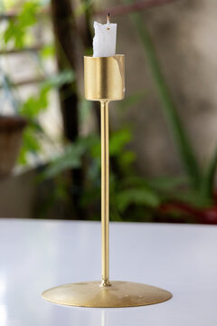 Modern tall golden metal candlestick holder with a white unlit candle placed on a table against blurred green natural background.