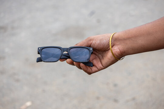 Close-up of a person holding dark matte black square sunglasses outdoors with a textured ground blurred background.