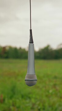 Vertical shot of audio microphone hanging upside down by cord in green meadow. Footage serves indie music festival promotion, audio podcast teaser and creative sound production campaign