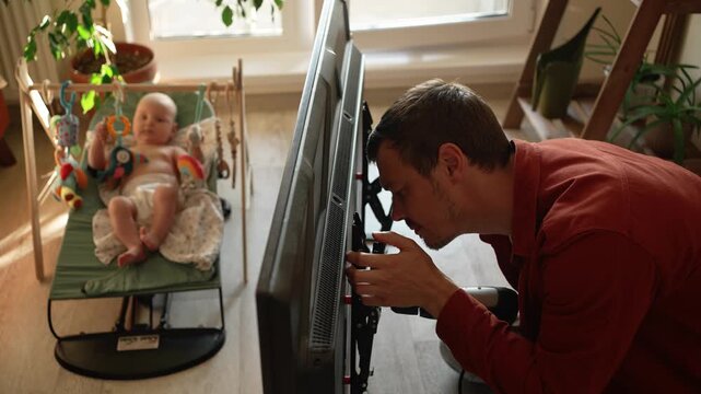 Young father assembling and installing a television wall bracket on a new tv in his living room, with his cute infant child observing from a baby bouncer nearby in the warm sunlight