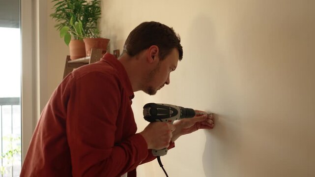 Concentrated man performing a diy home repair project, using a cordless power drill to make a hole in the plasterboard wall before blowing away the dust for a wall anchor