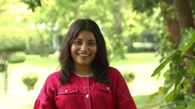 Happy indian Young adult Gen Z girl with looking at camera outdoors in summer park, concepts of joy, happiness, and a positive lifestyle.