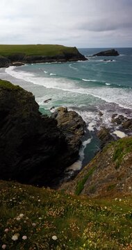 Waves breaking into a rocky cove beneath grassy cliffs in Cornwall, England, UK. Coastal wildflowers overlook the sea under a cloudy afternoon sky. Vertical han