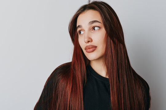 Young woman with long straight brown hair looking sideways with thoughtful expression. Casual portrait with natural makeup and plain background for beauty and lifestyle concept.