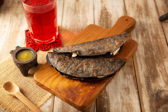 Mexican quesadillas made with blue corn tortillas filled with melted cheese and mushrooms served on a wooden cutting board and a glass of red hibiscus water (agua de jamaica) in the background.