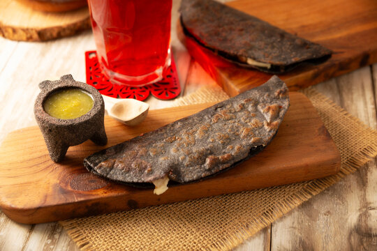 Mexican quesadillas made with blue corn tortillas filled with melted cheese and mushrooms served on a wooden cutting board and a glass of red hibiscus water (agua de jamaica) in the background.
