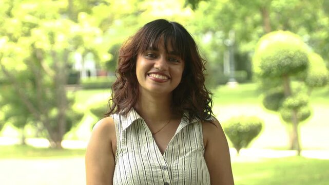 Happy indian Young adult Gen Z girl with looking at camera outdoors in summer park, concepts of joy, happiness, and a positive lifestyle.
