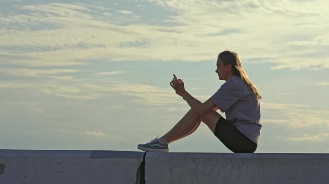 Side view of a beautiful young woman sitting alone on a concrete parapet against a cloudy sky, casually scrolling through social media on her smartphone during a peaceful sunset evening