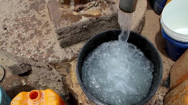 A close-up view of clear water pouring from a metal pipe into a black plastic bucket, with other containers visible nearby on the ground.