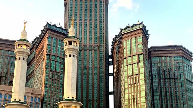 A low-angle view of the iconic Abraj Al Bait clock tower complex in Mecca, Saudi Arabia, set against a clear blue sky with light clouds.