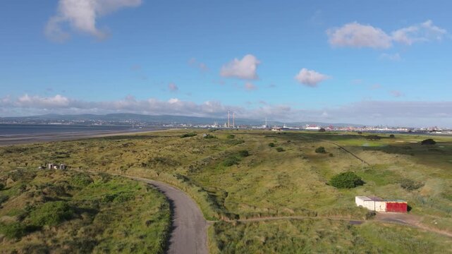 Curved access road winds through wild Irish grassland toward distant Poolbeg chimneys and tidal flats on a sunny day.