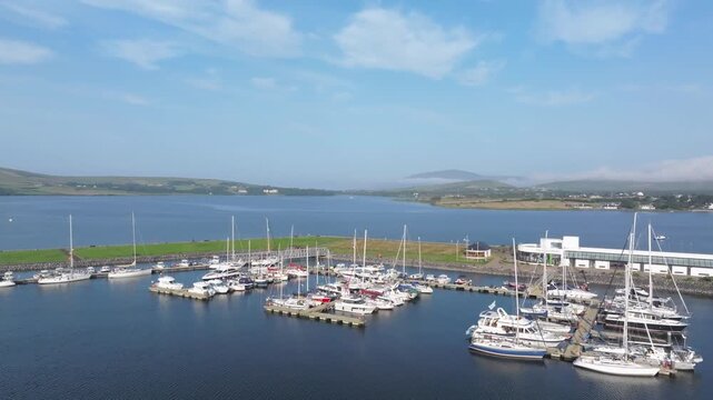 Yachts and motorboats fill a calm marina beside a wide coastal inlet as a drone glides overhead on a clear summer day.
