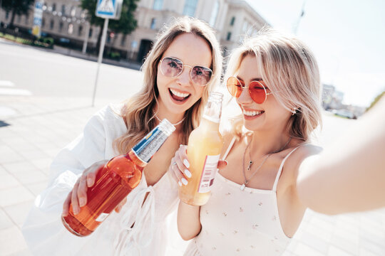 Two young beautiful smiling hipster female in trendy summer white dress. Carefree women posing outdoors. Positive models holding, drinking fresh cocktail drink in glass bottle. In the street, selfie