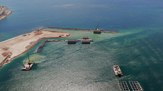 Exceptional aerial perspective of a major coastal engineering project, showing floating crane barges maneuvering and installing large concrete caissons to build a new port marina breakwater