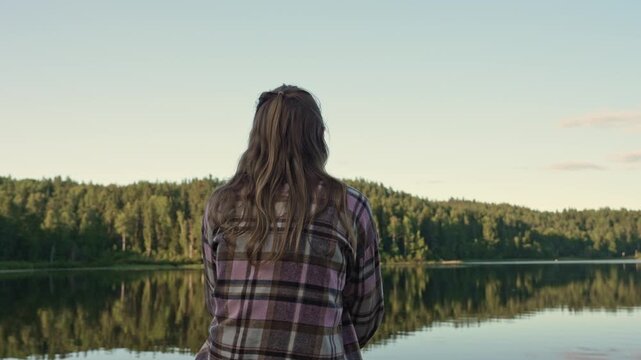 Solitary woman in a plaid shirt gazes across the calm waters of a mirror lake in Ontario as the soft golden light of evening descends upon the dense forest landscape during the quiet summer twilight.