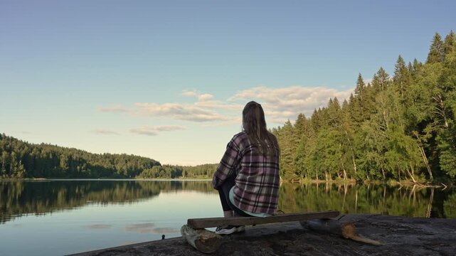 Serene woman sits peacefully on a weathered dock overlooking the reflective surface of a quiet forest lake during the golden hour as soft light touches the trees and gentle water in rural Sweden.