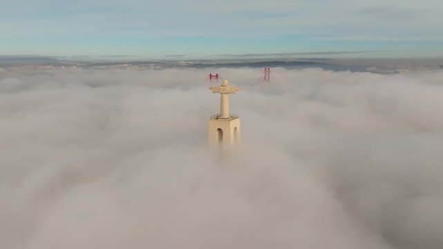 Slow pan drone shot of Cristo Rei statue, Lisbon at sunrise. Dense fog covers water. Sanctuary of Christ the King and 25 de Abril Bridge visible above mist. Almada, Portugal