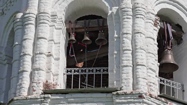 Golden sun illuminates the historic white stone belfry of the Holy Trinity Monastery as rhythmic bronze bells swing in harmony, creating a peaceful melody on a bright summer afternoon in Russia.