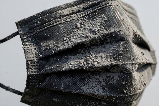 Close up of a disposable black medical face mask heavily covered in thick grey dust particles reflecting environmental pollution and waste issues