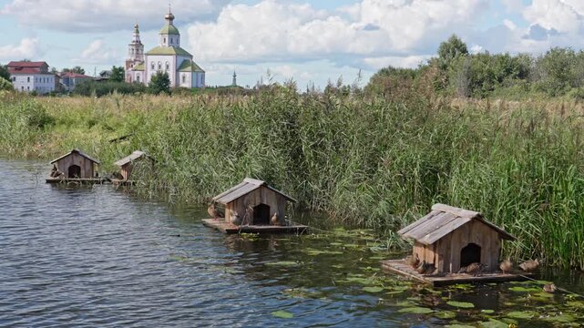 Small wooden homes drift gently upon the rippling river water as ducks swim past the golden reeds while the historic church towers under a bright blue sky on a calm and warm summer afternoon.