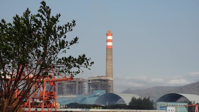 Industrial power plant facility with tall striped chimney stack and large storage domes against clear sky and mountain backdrop viewed through trees.