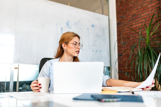 A young, professional businesswoman with red hair and glasses sits at a desk, holding a white coffee mug and reviewing business charts and graphs on a laptop. In a modern office.
