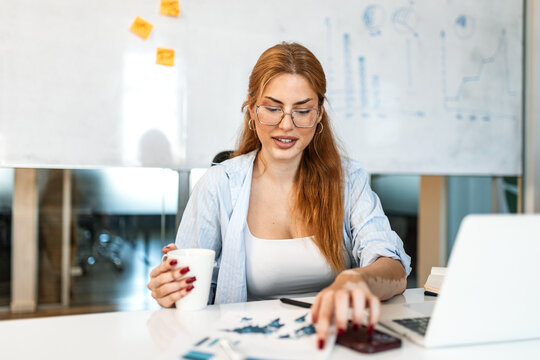 Professional businesswoman in her office with red hair and glasses, smiling happily while holding a white coffee mug. She works at a desk with a laptop, papers, and phone.